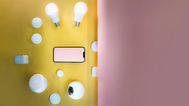Smart home tech gadgets including smart bulbs, Wi-Fi plug, security camera, and smartphone on a yellow background with pink accent wall for innovative travel and daily life solutions.